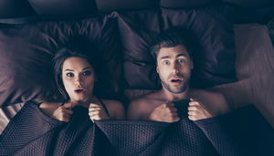 Funny lovers man and woman lying in bed covered by sheets with wide opened eyes and mouth looking at camera, having shocked and scared expressions, afraid to be found in bed by someone who unexpectedly came home from work